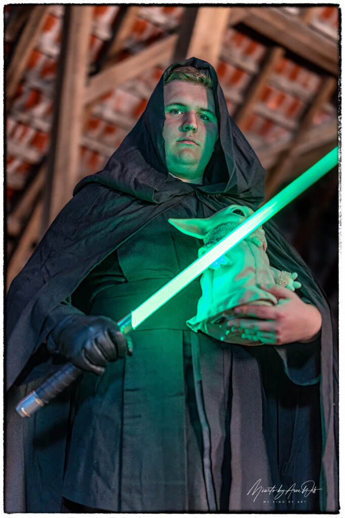Männlicher Cosplayer mit Jedi-Robe, Grogu im Arm und grün leuchtendem Lichtschwert.