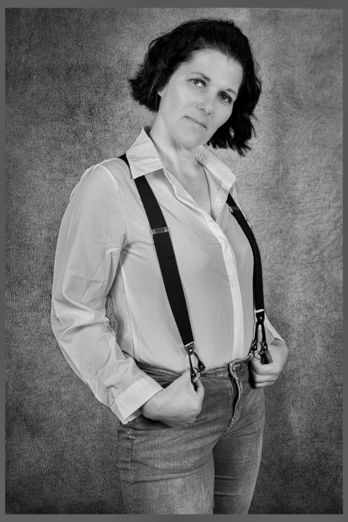 Selbstbewusste Frau im Schwarz-Weiß-Porträt mit offenem Hemd, Jeans und Hosenträgern – klassisch-cooler Look mit Retro-Flair.