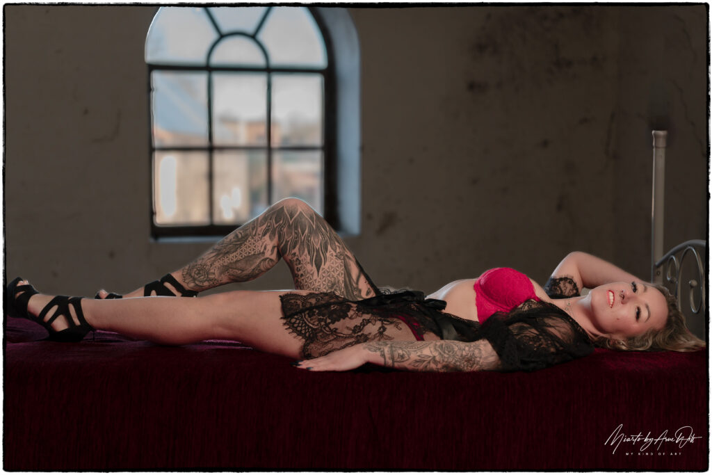 Frau liegt auf dem Bett vor einem Fenster â€“ dezente Pose in rotem Spitzen-BH.